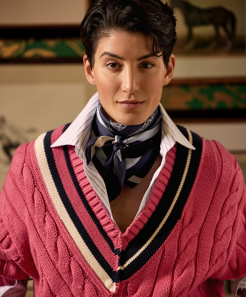 On left, A non-binary model wearing a sweater and silk neck scarf. On right, a male model wearing a white linen suit, a pink cable knit sweater, and a dress shirt with a tie.